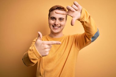 İzole edilmiş sarı arka planda rahat süveter giyen genç, kızıl saçlı adam gülümseyerek elleri ve parmakları gülen yüzlü bir çerçeve yapıyor. Yaratıcılık ve fotoğrafçılık kavramı.