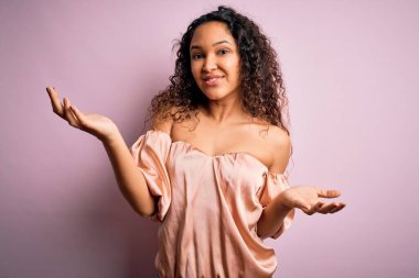 Kıvırcık saçlı, rahat tişörtlü, pembe arka planda gülümseyen, dostça kucaklaşan, olumlu ve kendinden emin genç bir kadın.