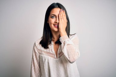 Mavi gözlü genç esmer kadın izole edilmiş beyaz arka plan üzerine günlük tişört giyiyor. Bir gözünü eliyle kapatıyor. Yüzünde kendine güvenen bir gülümseme ve şaşırtıcı bir duygu var..