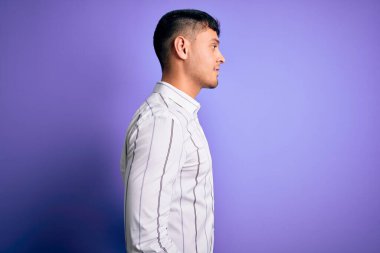 Zarif iş gömleği giyen yakışıklı genç adam mor arka planda duruyor. Yan tarafa bakıyor, rahat bir görünüm takınıyor ve kendine güvenen bir gülümseme sergiliyor..