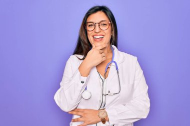 Mor arka planda steteskop ve tıbbi ceket giyen profesyonel doktor kadın kameraya çapraz kollar ve çeneyle gülümseyerek bakıyor. Olumlu düşünüyorum..