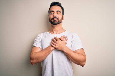 Sakallı, sıradan tişörtlü, beyaz arka planda duran, elleri göğsünde, gözleri kapalı ve yüzünde minnet ifadesi olan genç yakışıklı bir adam. Sağlık kavramı.