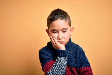 Sarı arka plan üzerine kışlık süveter giyen küçük bir çocuk kollarını kavuşturarak depresyondan sıkılmış ve yorgun göründüğünü düşünüyor..