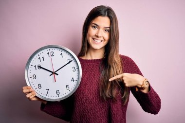 Geri sayım yapan genç ve güzel bir kız, pembe arka planda büyük bir saat tutuyor. Şaşırmış yüz ifadesi kendini gösteriyor.