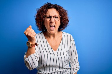 Orta yaşlı güzel kıvırcık saçlı kadın izole edilmiş arka planda rahat çizgili tişört giyiyor kızgın ve kızgın yumruğunu kaldırıyor ve öfkeyle bağırıyor. Öfke ve agresif kavram.