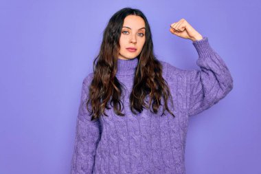 Genç, güzel bir kadın, balıkçı yaka kazak giyiyor mor arka planda dikiliyor güçlü biri kol kası gösteriyor, kendinden emin ve gururlu.