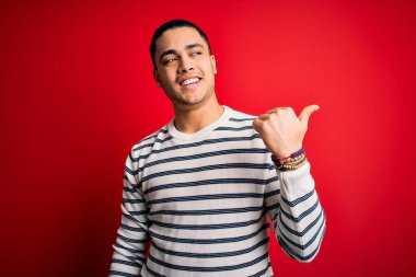 İzole edilmiş kırmızı arka planda dikilen sıradan çizgili tişört giyen genç Brezilyalı adam gülümsüyor ve başparmağıyla yana bakıyor..