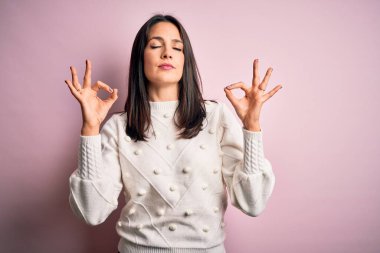 Mavi gözlü genç esmer kadın izole edilmiş pembe arka planda gündelik süveter giyiyor rahatlıyor ve parmaklarıyla meditasyon hareketi yaparken gözleri kapalı gülümsüyor. Yoga konsepti.