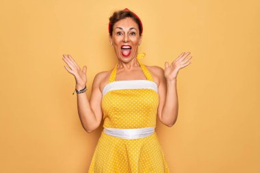 Orta yaşlı bir kadın, 50 'li yaşlarda sarı arka plan üzerine eski tarz elbise giyip çılgın ve başarıyı kutlarken kollarını kaldırmış ve gözleri açık bir şekilde heyecanla haykırıyor. Kazanan konsept