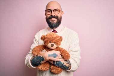 Sakallı ve dövmeli yakışıklı kel bir adam ayıcığı soyutlanmış pembe arka planda tutuyor. Mutlu bir yüzü var ve gülümsüyor.