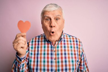 Kıdemli yakışıklı, ağarmış romantik bir adam pembe arka planda kırmızı kağıt kalp şeklinde bir kalp tutuyor. Şaşırmış bir yüzle korkmuş ve korku ifadesinden heyecanlanmış.