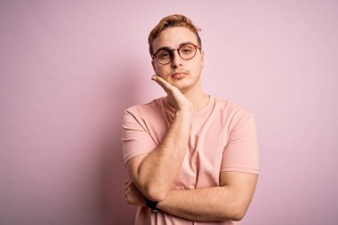 Genç, kızıl saçlı, günlük tişört giyen, izole edilmiş pembe arka planda duran, depresyondan sıkılmış ve yorgun görünen, kollarını kavuşturmuş genç bir adam..