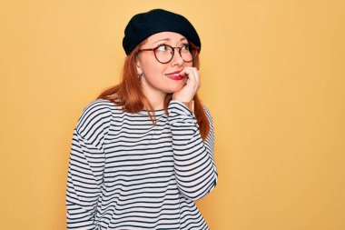 Sarı arka planda çizgili tişört ve Fransız beresi giyen güzel kızıl saçlı kadın elleri ağız yiyen tırnaklarla gergin ve stresli görünüyor. Endişe sorunu.