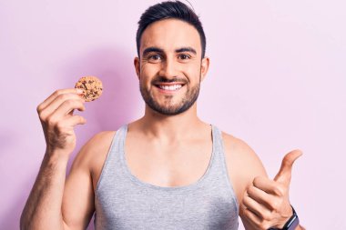 Sakallı, kolsuz tişörtlü, çikolatalı kurabiye yiyen mutlu ve pozitif gülümseyen, başparmağını kaldıran mükemmel ve onay işareti yapan genç yakışıklı bir adam.