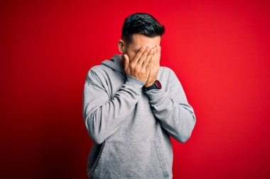 Eşofman giyen genç yakışıklı sporcu, izole edilmiş kırmızı arka planda dikiliyor. Ağlarken suratını ellerle kapatıyor. Depresyon kavramı.