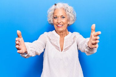 Son sınıftaki gri saçlı kadın, günlük kıyafetler giyip kameraya bakıyor ve sarılmak için kollarını açmış gülümsüyor. Mutluluğu kucaklayan neşeli bir ifade.. 