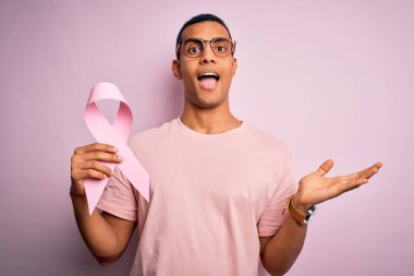 Genç ve yakışıklı Afro-Amerikalı adam pembe kanser kurdelesini izole edilmiş arka planda tutuyor çok mutlu ve heyecanlı, kazanan yüz ifadesi zaferi büyük bir gülümsemeyle kutluyor ve ellerini kaldırıyor.