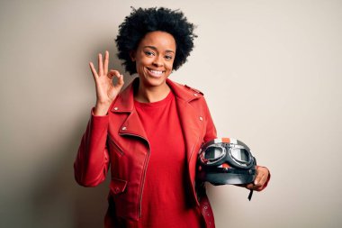 Kıvırcık saçlı, motosiklet kaskı tutan ve gülümseyen Afro-Amerikan motosikletli genç bir kadın el ve parmaklarıyla tamam işareti yapıyor. Başarılı ifade.