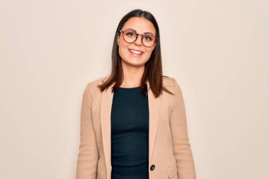 Güzel, esmer bir iş kadını beyaz arka planda ceket ve gözlük takıyor ve yüzünde mutlu ve havalı bir gülümseme var. Şanslı kişi..