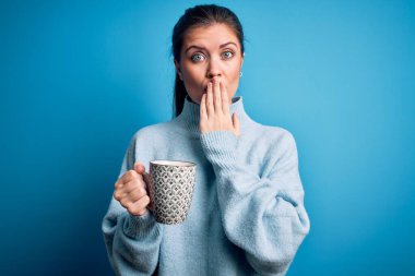 Mavi gözlü genç bir kadın izole edilmiş arka planda kahve fincanı içerek ağzını kapatıyor. Utanç verici bir hata, korku ifadesi, sessizlikten korkmak, gizli bir konsept.