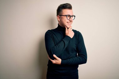 Gözlük takan yakışıklı beyaz bir adam ve izole edilmiş arka planda, elleri çenesinde soru soran, düşünceli bir yüz ifadesi olan sıradan bir süveter giyiyor. Düşünceli bir yüzle gülümsüyor. Şüphe kavramı.