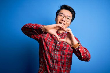 Mavi arka plan üzerinde gözlük takan genç, yakışıklı Çinli bir adam elleriyle kalp sembolü yapan bir aşkla gülümsüyor. Romantik konsept.