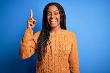 Genç Afro-Amerikalı kadın günlük sarı kazak giyiyor mavi izole arka planda duruyor ve başarılı bir fikri işaret ediyor. Heyecanlı ve mutlu. Bir numara..
