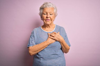 Son sınıf güzeli, gri saçlı kadın, pembe arka plan üzerine günlük tişört giymiş, elleri göğsünde, gözleri kapalı ve yüzünde minnet dolu bir jest ile gülümsüyor. Sağlık kavramı.