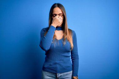 Mavi gözlüklü, genç, sarışın bir kadın mavi arka planda dikiliyor. Burnu ve gözleri ovuşturuyor, yorgunluk ve baş ağrısı çekiyor. Stres ve hüsran kavramı.