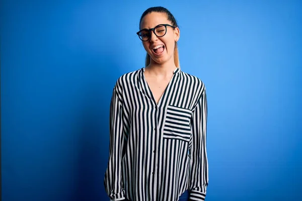 Beautiful blonde woman with blue eyes wearing striped shirt and glasses over blue background winking looking at the camera with sexy expression, cheerful and happy face.
