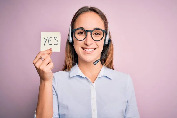 Beautiful call center agent woman working using headset holding reminder with yes message with a happy face standing and smiling with a confident smile showing teeth