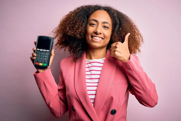 Young african american woman with afro hair holding dataphone over pink background happy with big smile doing ok sign, thumb up with fingers, excellent sign