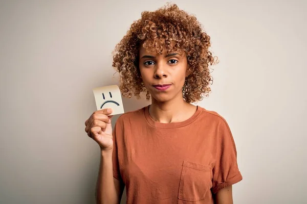 Young african american woman with curly hair holding reminder paper with sad face emoji with a confident expression on smart face thinking serious