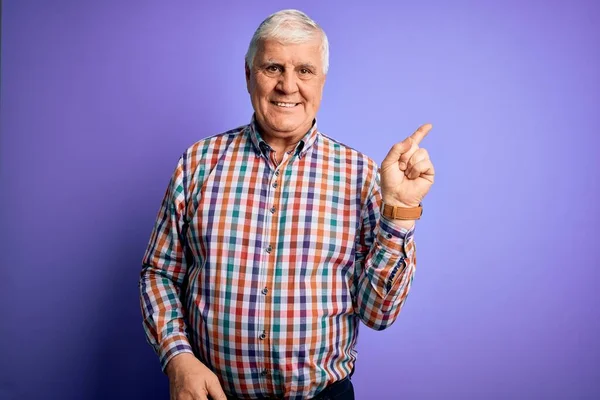 stock image Senior handsome hoary man wearing casual colorful shirt over isolated purple background with a big smile on face, pointing with hand finger to the side looking at the camera.