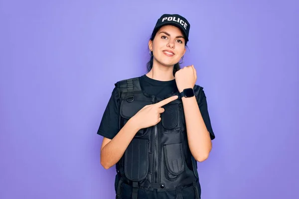 Young police woman wearing security bulletproof vest uniform over purple background In hurry pointing to watch time, impatience, looking at the camera with relaxed expression