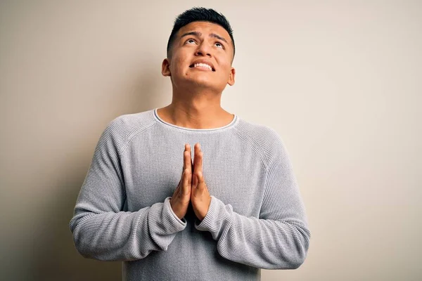 Young handsome latin man wearing casual sweater standing over isolated white background begging and praying with hands together with hope expression on face very emotional and worried. Begging.