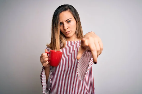 Beautiful blonde woman with blue eyes drinking red mug of coffee over white background pointing with finger to the camera and to you, hand sign, positive and confident gesture from the front