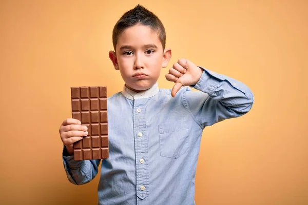 若い男の子食べる甘いチョコレートバーのためのデザート上の孤立した黄色の背景で怒っている顔 否定的な兆候を示す嫌いで親指ダウン 拒否の概念 — ストック写真