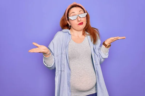 Young redhead pregnant woman expecting baby wearing funny thug life sunglasses clueless and confused expression with arms and hands raised. Doubt concept.