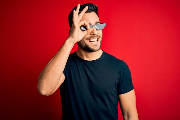 Young handsome man wearing funny thug life sunglasses over isolated red background doing ok gesture with hand smiling, eye looking through fingers with happy face.