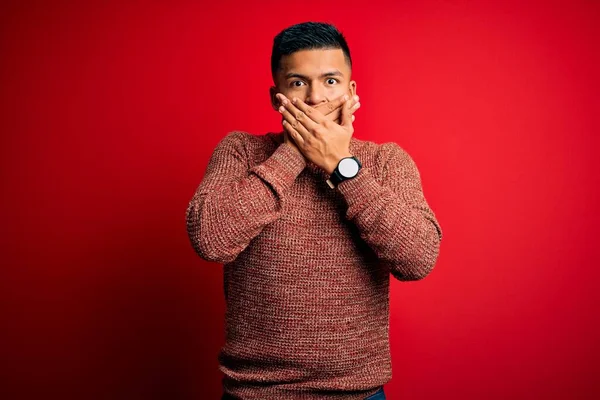 Young handsome latin man wearing casual sweater standing over red background shocked covering mouth with hands for mistake. Secret concept.