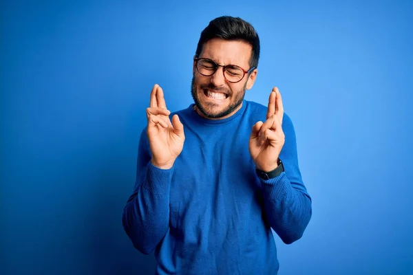 Young handsome man with beard wearing casual sweater and glasses over blue background gesturing finger crossed smiling with hope and eyes closed. Luck and superstitious concept.