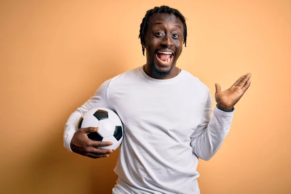 Young african american player man playing soccer holding football ball over yellow background very happy and excited, winner expression celebrating victory screaming with big smile and raised hands