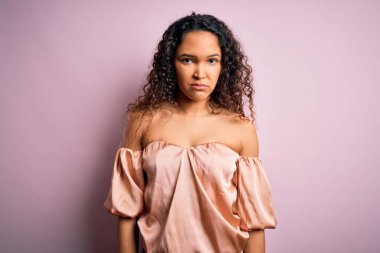 Kıvırcık saçlı, rahat tişörtlü, pembe arka planda duran, kuşkucu ve gergin, sorunlu, genç ve güzel bir kadın. Negatif kişi.