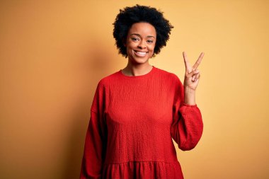 Kıvırcık saçlı, sıradan süveter giyen, kameraya gülümseyen ve zafer işareti yapan güzel Afro-Amerikan kadın. İki numara..