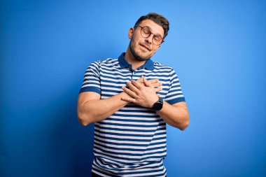 Mavi gözlü, gözlüklü ve mavi arka planda sıradan çizgili tişörtlü elleri göğsünde, gözleri kapalı ve yüzünde minnet dolu bir jest olan genç bir adam. Sağlık kavramı.