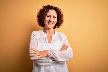 Orta yaşlı güzel kıvırcık saçlı kadın sarı arka planda rahat bir yaz elbisesi giyiyor ve kollarını kavuşturup kameraya bakıyor. Pozitif kişi.