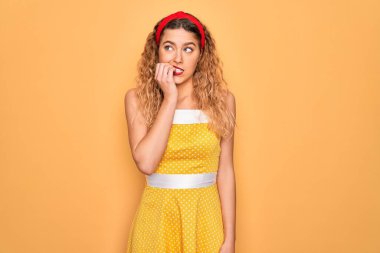 Sarı arka planda dikiliyken mavi gözlü güzel sarışın kadın elleri ağızda tırnak yerken gergin ve stresli görünüyor. Endişe sorunu.