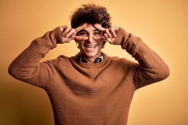 İzole edilmiş sarı arka plan üzerine günlük gömlek ve süveter giyen genç yakışıklı adam barış sembolü yapıyor. Yüzünde parmaklar, gülümseyerek zaferi gösteriyor.