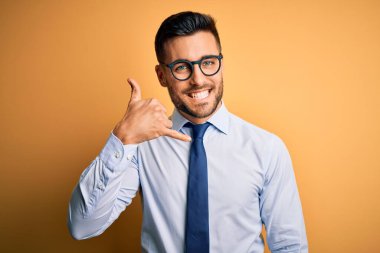 Kravat takan ve sarı arka planda duran gözlüklü genç yakışıklı işadamı telefonla el ve parmaklarıyla telefonla konuşuyor gibi gülümsüyor. İletişim kavramları.